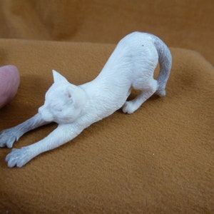 Könnte beinhalten: Weiße Keramik-Katzenfigur mit grauem Schwanz, die ihre Vorderpfoten ausstreckt. Die Figur steht auf einer braunen Oberfläche.