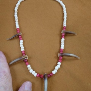 Peut inclure: Un collier fait à la main composé d'un fil de perles blanches et rouges, orné de plusieurs pendentifs en forme de griffes brunes. Le collier est fermé par un fermoir doré. Les perles sont petites et rondes, créant une esthétique rustique et naturelle.