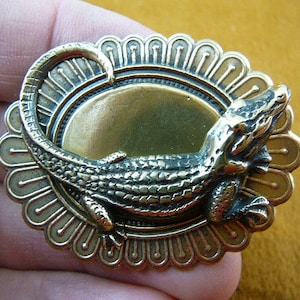 May include: A gold-toned alligator brooch with a detailed, textured design. The alligator is positioned on a circular, ornate base with a raised, circular centre.