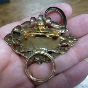 May include: A gold-toned brooch with an ornate design. The brooch has a ring on the back for hanging.