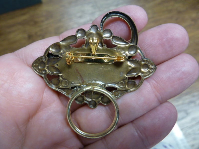 May include: A gold-toned brooch with an ornate design. The brooch has a ring on the back for hanging.