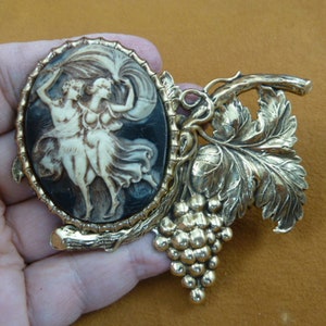 May include: A gold-tone brooch with a cameo of two women in a grapevine design. The cameo is set in an oval frame and the grapevine design features leaves and grapes.