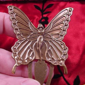 Könnte beinhalten: Ein bronzefarbener Schmetterlingshaarkamm mit einem detaillierten Design einer Fee mit Flügeln. Der Kamm hat ein dekoratives Design mit einer erhabenen Textur.