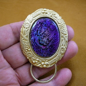 May include: A gold-toned key chain with an oval-shaped abalone shell inlay. The shell has a purple and iridescent colour. The key chain has a floral design around the shell.