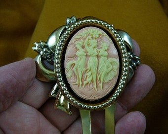 May include: A gold-tone hair clip with a cameo featuring three women in a pink and yellow design.