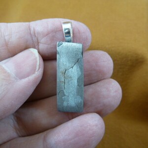 May include: A silver pendant with a rectangular piece of meteorite. The meteorite has a gray color and a rough, textured surface with visible cracks.