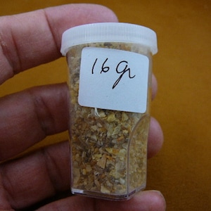 May include: A small, clear plastic jar with a white lid, filled with a tan, granular substance. A white label on the jar reads "16g" in black ink. The jar is held in a hand against a blurred, yellow-brown background.