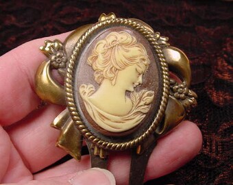 May include: A gold-tone hair comb featuring a cameo of a woman's profile. The cameo is set in a round frame with a rope-like border. The comb has a bow-shaped design on either side of the cameo.