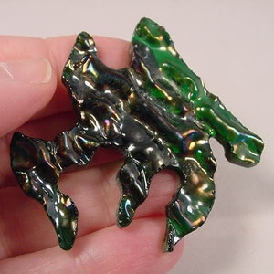 Op de afbeelding: Een groene en zwarte iriserende glazen hanger in de vorm van een abstract ontwerp. De hanger heeft een golvende, organische vorm met een gat in het midden.