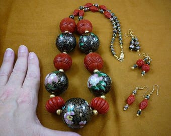 Puede incluir: Un collar de cuentas negras y rojas con diseños florales y pendientes a juego. El collar está hecho de cuentas redondas grandes con esmalte negro y patrones florales en blanco y rosa. Los pendientes también son redondos y presentan los mismos diseños florales.