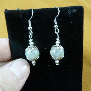 Peut inclure: Une paire de boucles d'oreilles pendantes avec des crochets et des accents en métal argenté. Chaque boucle d'oreille présente une pierre ronde vert pâle sertie dans un décor argenté en filigrane. Les boucles sont présentées sur fond noir.