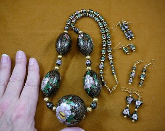 Puede incluir: Un collar negro y dorado con cuentas de esmalte floral y pendientes a juego. El collar tiene una larga cadena de cuentas con una cuenta más grande de forma ovalada en el centro. Los pendientes son pequeños y colgantes.