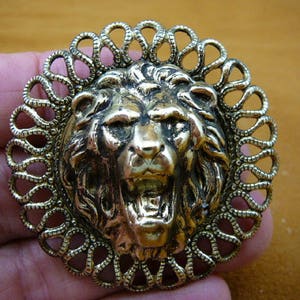 Puede incluir: Un broche de cabeza de león dorado con un diseño detallado. La melena del león está esculpida de forma intrincada y la cabeza está rodeada por un marco circular decorativo.