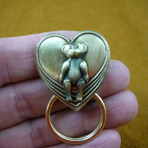 以下が含まれることがあります： 小さなテディベアチャームが入った、ゴールドトーンのハート型のキーチェーン。ハートは装飾的なボーダーと、下部にリングが付いています。