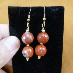 May include: A pair of dangle earrings featuring two faceted, reddish-brown stone beads stacked on gold-colored metal findings. The top bead has a lighter, mottled pattern. The earrings are displayed on a black velvet surface.