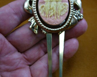May include: A gold hair comb with a cameo featuring three women in a pink and white design. The cameo is set in a gold oval frame with a beaded edge.