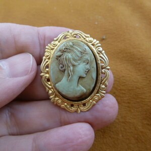 May include: A gold-tone cameo brooch featuring a white cameo of a woman's profile.