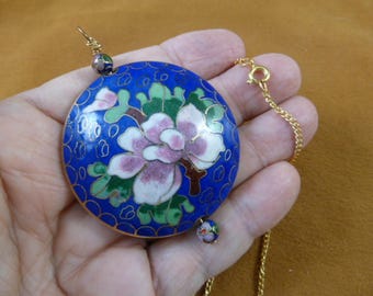 Könnte beinhalten: Ein blauer und weißer Cloisonné-Anhänger mit floralem Design hängt an einer goldenen Kette. Der Anhänger ist rund und zeigt eine weiße Blume mit rosa Akzenten, umgeben von grünen Blättern. Die Kette besteht aus kleinen goldenen Gliedern.