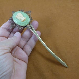 May include: A green and gold hair stick with a cameo of a woman's profile. The stick is about 7 inches long and has a delicate filigree design.