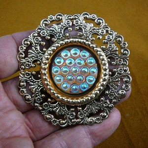 Könnte beinhalten: Eine goldfarbene Brosche aus Metall mit einem runden Design. Die Mitte der Brosche zeigt ein rundes, irisierendes blaues Glasstück mit einem Muster aus kleinen Kreisen.