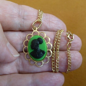 May include: Gold-coloured necklace with a cameo pendant. The pendant has a black silhouette on a green background, set in a scalloped frame. The chain is a delicate gold colour.