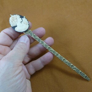 May include: A gold hair stick with a black cameo featuring a woman's profile. The stick is decorated with a floral design.