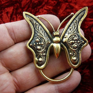 Könnte beinhalten: Eine goldfarbene Schmetterlingsbrosche mit filigranen Details und schwarzem Hintergrund. Der Schmetterling hat einen abgerundeten Körper und zarte Flügel.