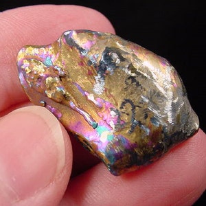 May include: A close-up of a small, iridescent, metallic rock with a rough, textured surface. The rock has a variety of colors, including gold, purple, and blue.