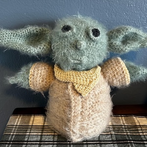 K&ouml;nnte beinhalten: Ein geh&auml;keltes Pl&uuml;schtier, das einem Charakter aus dem Star Wars-Franchise &auml;hnelt. Das Spielzeug ist gr&uuml;n und braun mit gro&szlig;en schwarzen Augen und einem gelben Kragen.