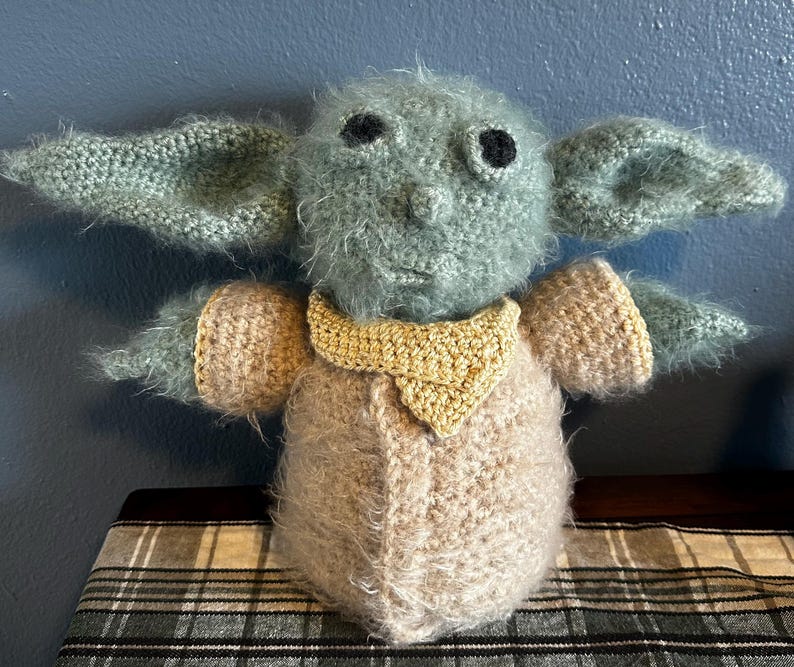 K&ouml;nnte beinhalten: Ein geh&auml;keltes Pl&uuml;schtier, das einem Charakter aus dem Star Wars-Franchise &auml;hnelt. Das Spielzeug ist gr&uuml;n und braun mit gro&szlig;en schwarzen Augen und einem gelben Kragen.