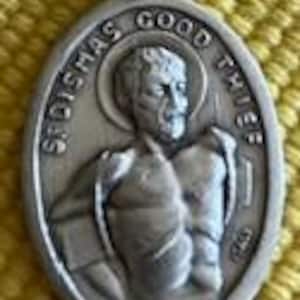 Może przedstawiać: Owalny, srebrzysty wisiorek z reliefem przedstawiającym mężczyznę z napisem "ST. DISMAS GOOD THIEF". Mężczyzna jest przedstawiony z aureolą. Wisiorek ma małą pętlę u góry do łańcuszka.