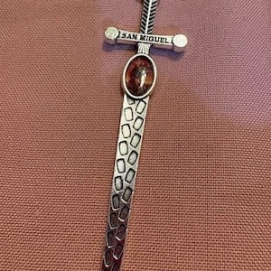 May include: Silver-toned sword pendant with the inscription "SAN MIGUEL" on the crossguard. The pendant features an oval-shaped, reddish-brown stone and a textured blade design. A small loop is at the top for hanging.