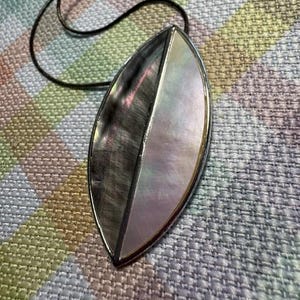 May include: A marquise-shaped pendant necklace featuring a split design. One half is composed of dark, textured grey shell, while the other half is iridescent, pearlescent white shell. The pendant is framed in metal and hangs from a thin black cord.