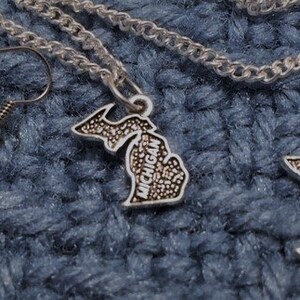 May include: A silver necklace and earrings set featuring a Michigan state outline pendant and earrings. The pendant and earrings have the word "Michigan" inscribed on them.