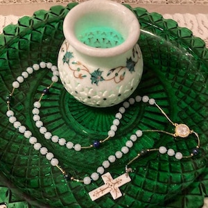 May include: A white rosary necklace with a white cross pendant and blue beads. The rosary is on a green plate with a white carved bowl.