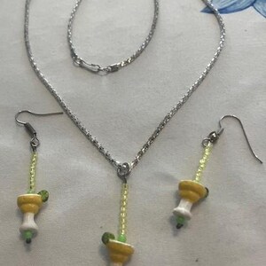 May include: A silver-toned necklace and earring set. The necklace has a chain with a margarita pendant. The earrings also feature margarita charms. The margarita charms are yellow, white, and green.