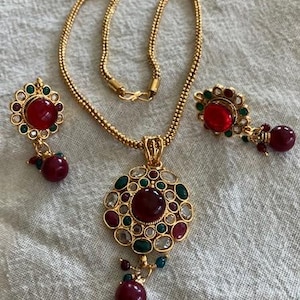 May include: A gold necklace with a pendant featuring a red stone surrounded by smaller green and white stones. The necklace comes with matching earrings with red stones.