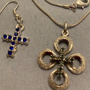 May include: A silver-toned jewelry set featuring a cross pendant necklace and a cross earring. The cross pendant has a four-leaf clover design with green gemstones. The earring is a cross shape with blue gemstones.