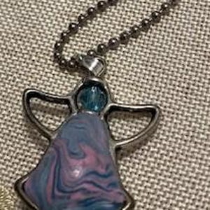 Puede incluir: Un collar con un colgante de &aacute;ngel de plata con una incrustaci&oacute;n de vidrio marmoleado azul, rosa y blanco. La cadena es de plata y tiene un dise&ntilde;o de eslabones de bola.