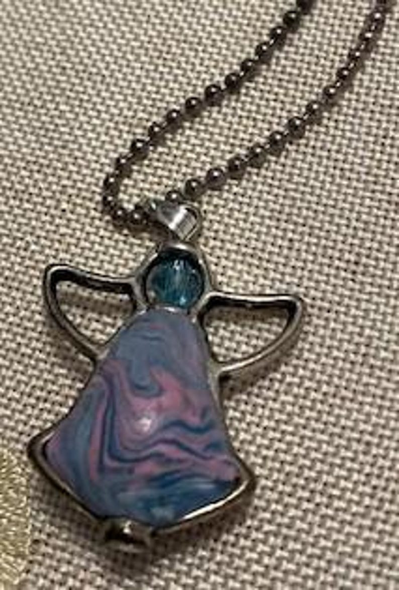 Puede incluir: Un collar con un colgante de &aacute;ngel de plata con una incrustaci&oacute;n de vidrio marmoleado azul, rosa y blanco. La cadena es de plata y tiene un dise&ntilde;o de eslabones de bola.