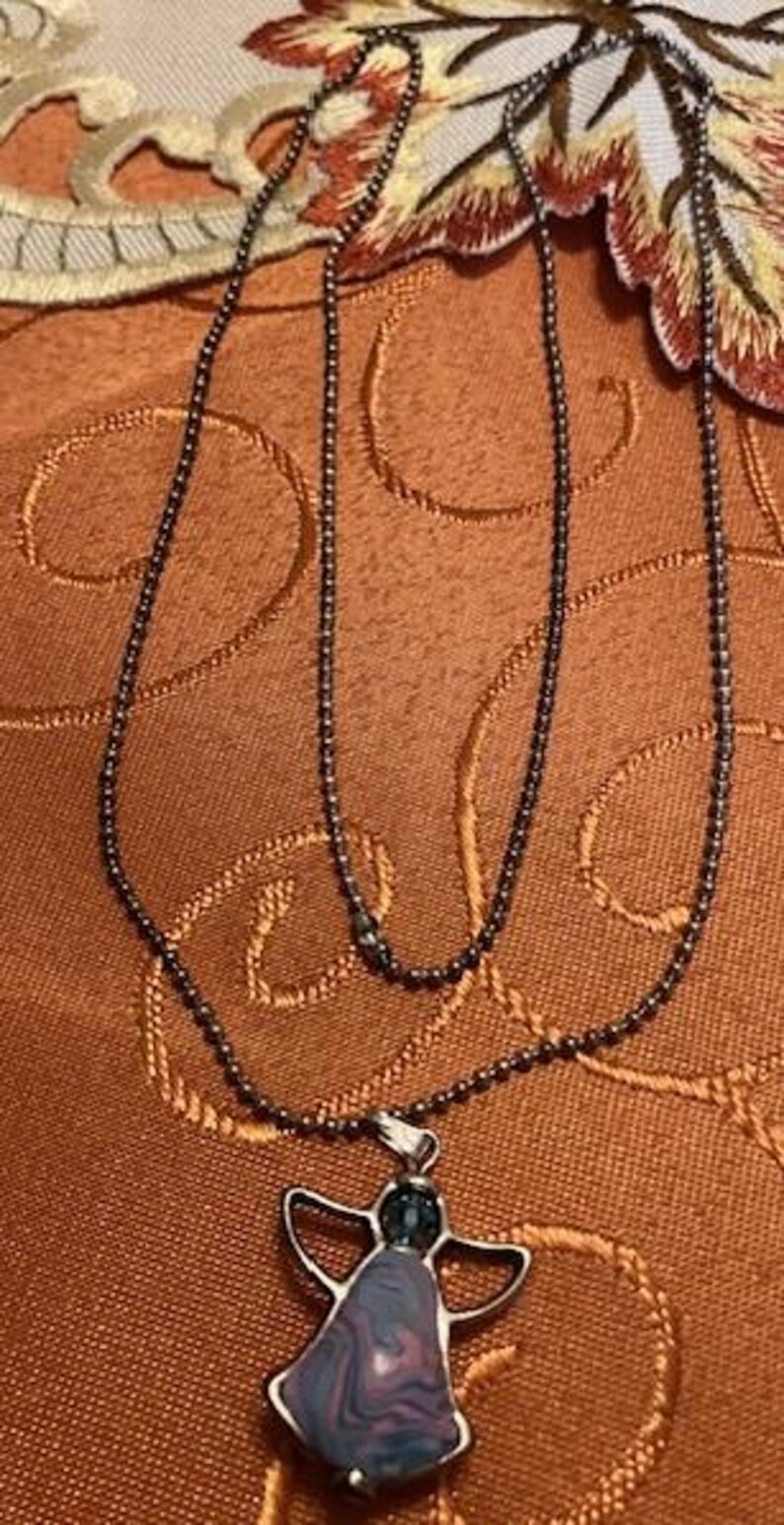 Puede incluir: Un collar de cadena de plata con un colgante de &aacute;ngel morado y azul. El &aacute;ngel est&aacute; hecho de una piedra con un patr&oacute;n de remolino.