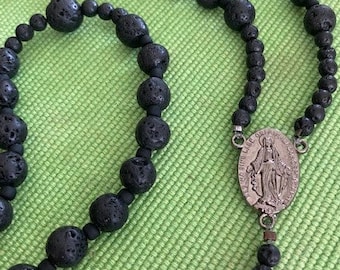 Puede incluir: Rosario de cuentas de lava negra con un colgante de plata que representa a la Virgen María. Las cuentas están enhebradas en un cordón negro.