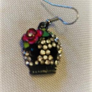 May include: A single, ornate earring featuring a black skull design adorned with sparkling crystals. A red rose with green leaves sits atop the skull. The earring has a silver-colored hook.