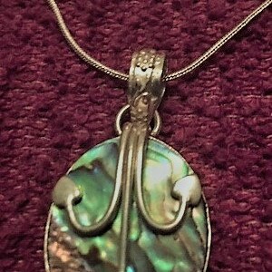 May include: A silver chain necklace with an oval abalone shell pendant. The pendant has a silver design in the shape of a stylized tree.