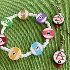 May include: A colorful bracelet with white beads and eight round charms featuring billiard ball numbers. The bracelet is paired with a set of earrings with a similar design, featuring a red triangle and a black eight ball.
