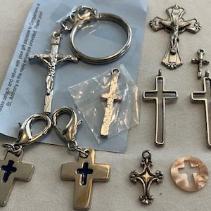 Op de afbeelding: Een verzameling zilverkleurige religieuze kruisen, waaronder kruisbeelden, hangerkruisen en sleutelhangers. Sommige kruisen hebben blauwe emaille accenten. De items variëren in grootte en ontwerp, geschikt voor sieraden of religieuze ambachten.