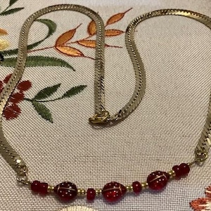 May include: A gold chain necklace with a row of red glass beads. The beads are oval shaped and have a slight shimmer.