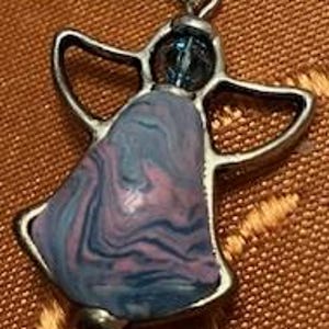 Puede incluir: Un colgante de &aacute;ngel de plata con una piedra de m&aacute;rmol azul y rosa. La piedra tiene un patr&oacute;n de remolino de azul, rosa y blanco.