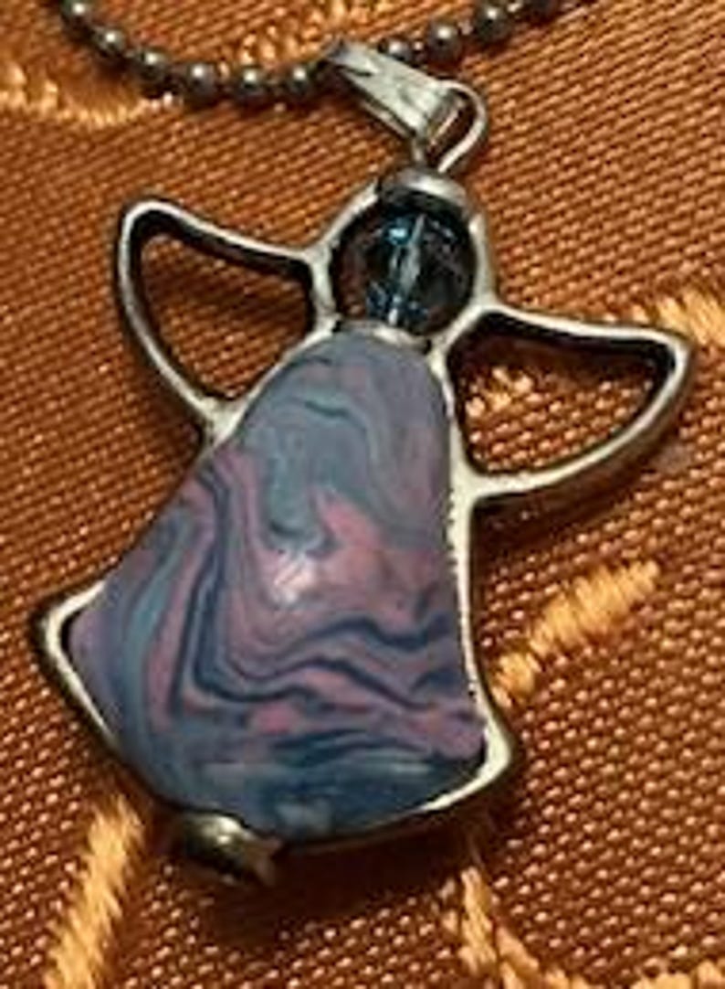 Puede incluir: Un colgante de &aacute;ngel de plata con una piedra de m&aacute;rmol azul y rosa. La piedra tiene un patr&oacute;n de remolino de azul, rosa y blanco.