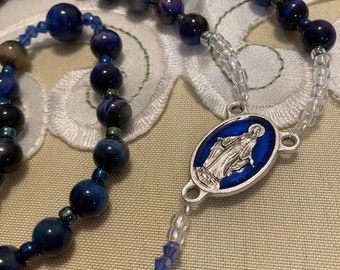 Può includere: Un rosario blu e argento con un ciondolo blu e argento che presenta un'immagine religiosa. Le perle sono fatte di pietra di occhio di tigre.